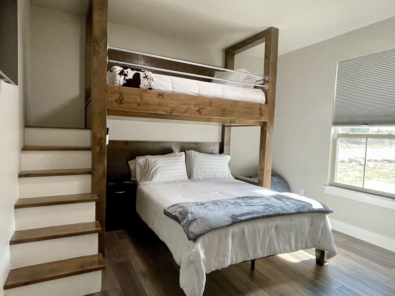 A bedroom with a wooden staircase leading to a top bunk above a made bed with white and gray bedding