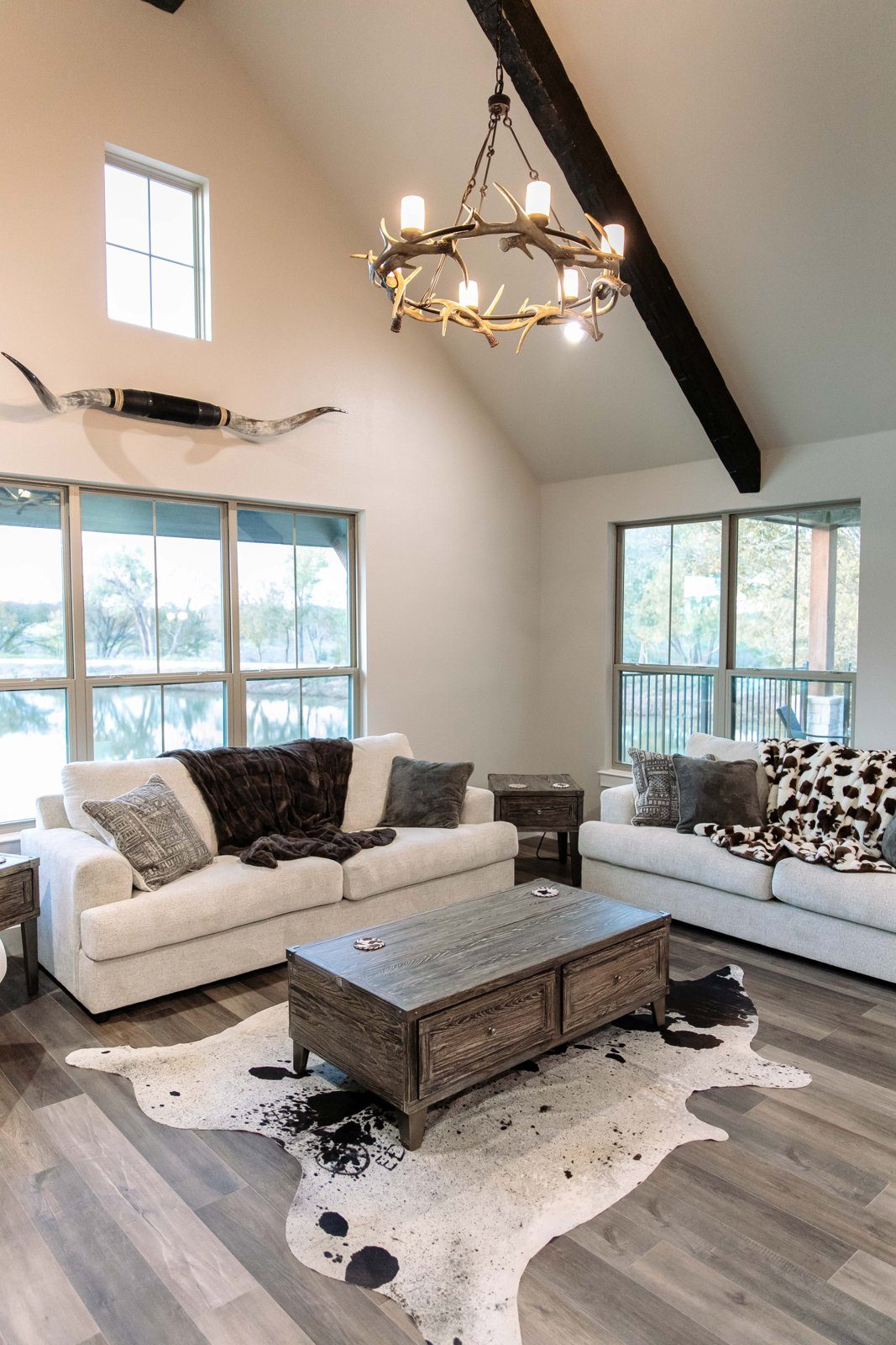 A villa living room with a vaulted white ceiling, dark beam, antler chandelier, mounted Longhorn horns, white sofa and cowhide rug